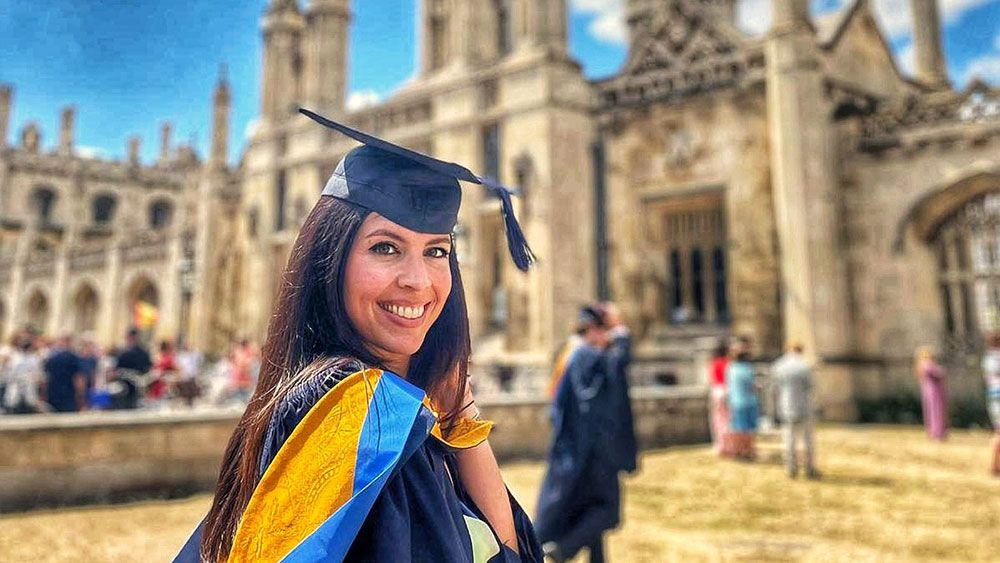 Smiling woman in cap and gown with blue and gold sash in front of university building.