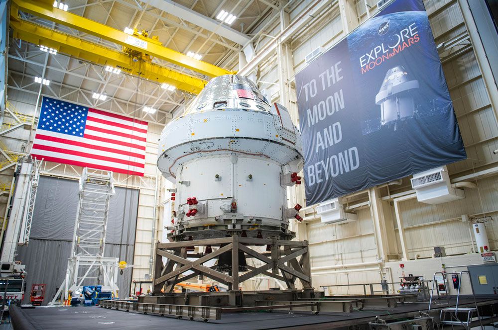 미국 국기가 한쪽 벽면에 있고 다른 쪽 벽면에 'To the Moon and Beyond'라는 배너가 있는 대형 시설 안에 있는 Orion 우주선.